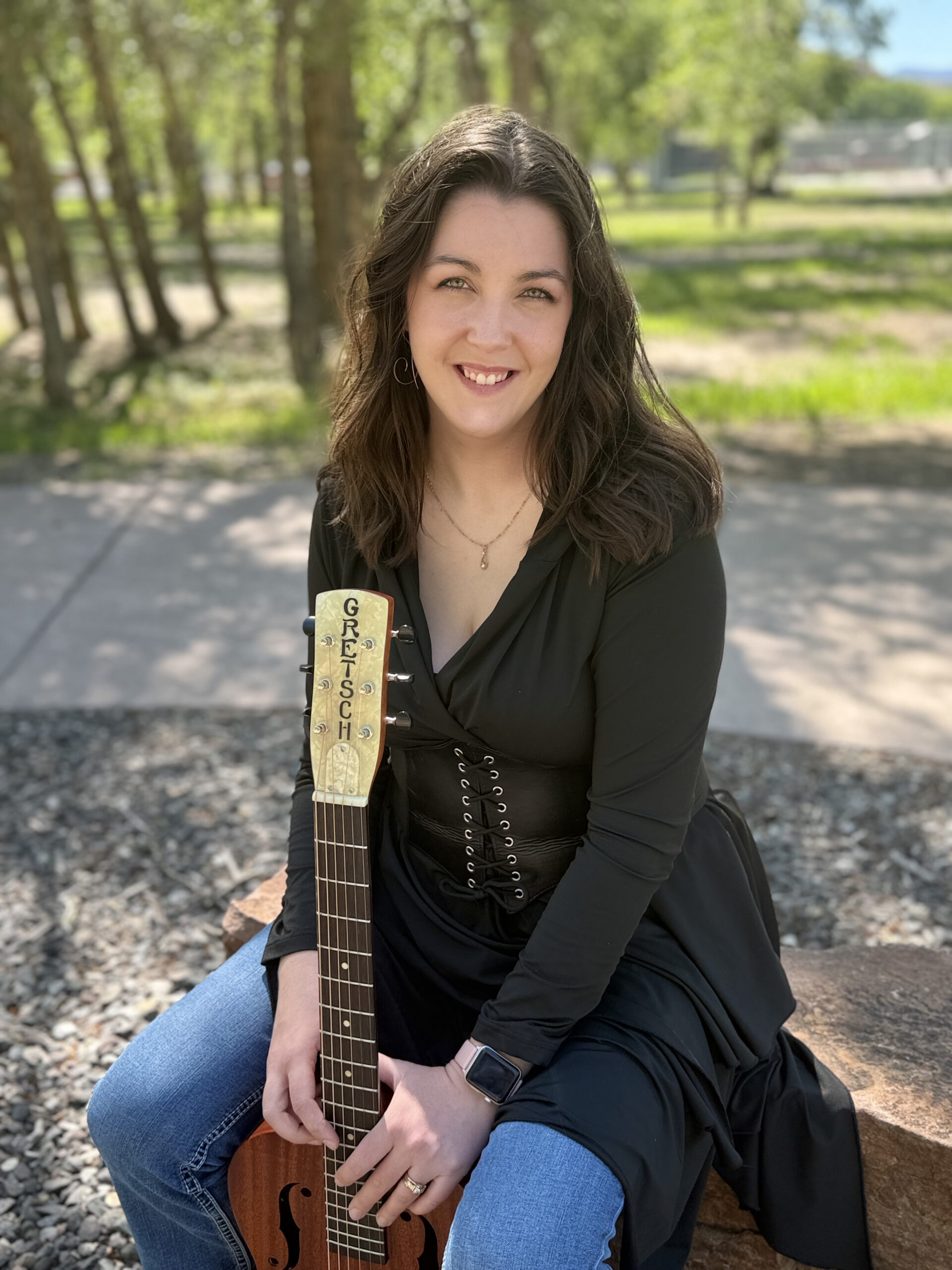 Image is of Jacinda Wilson smiling at the camera while loosely holding her guitar, dressed in a loose black top and jeans, with green grass and trees in the top half of the background and gray gravel in the lower half, split by a gray cement pathway in the middle. She is sitting on a large, reddish brown rock.